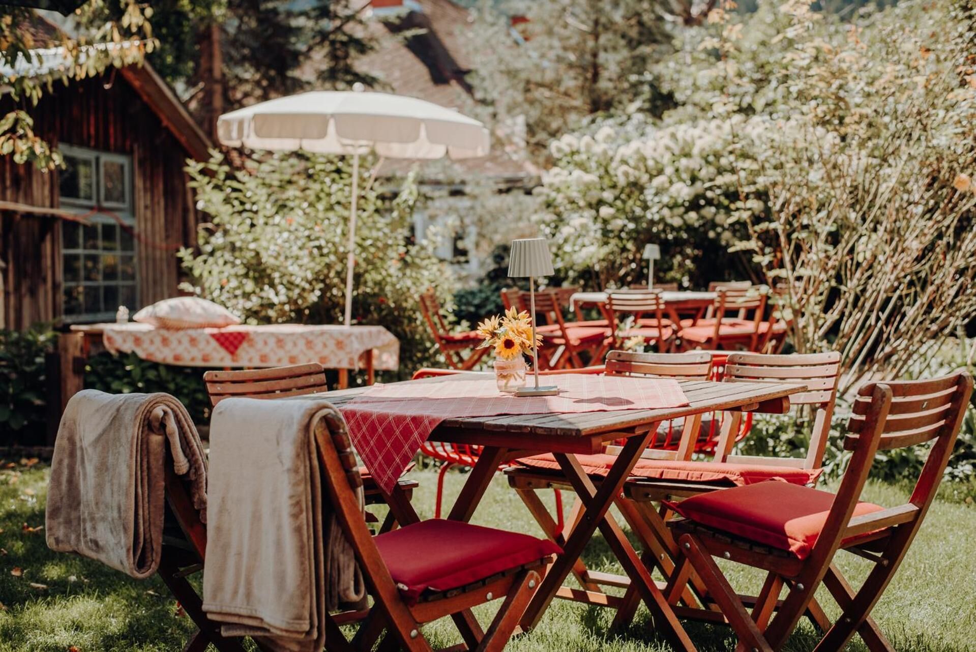 Un área de comedor al aire libre con mesas y sillas de madera, un paraguas blanco y un edificio de madera rústica al fondo.