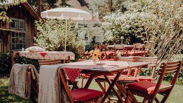 Un área de comedor al aire libre con mesas y sillas de madera, un paraguas blanco y un edificio de madera rústica al fondo.