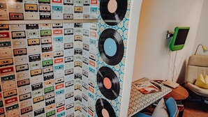 A room with a wall covered in cassette tapes, a wall-mounted record player, and a small table with a book and a lamp.