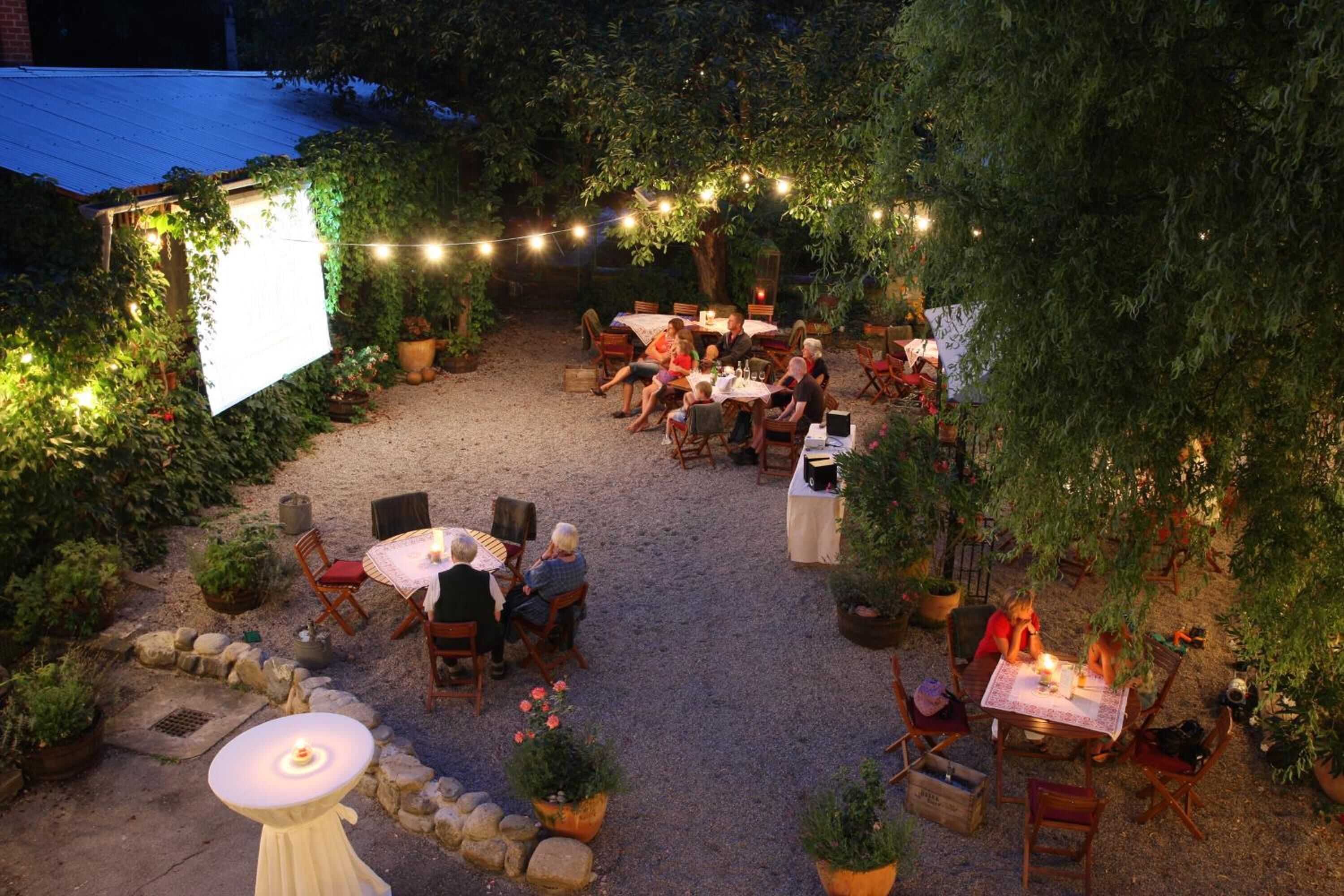 Eine Terrassenanlage im Freien mit mehreren Tischen und Stühlen, umgeben von Blumentöpfen und Lichterketten.
