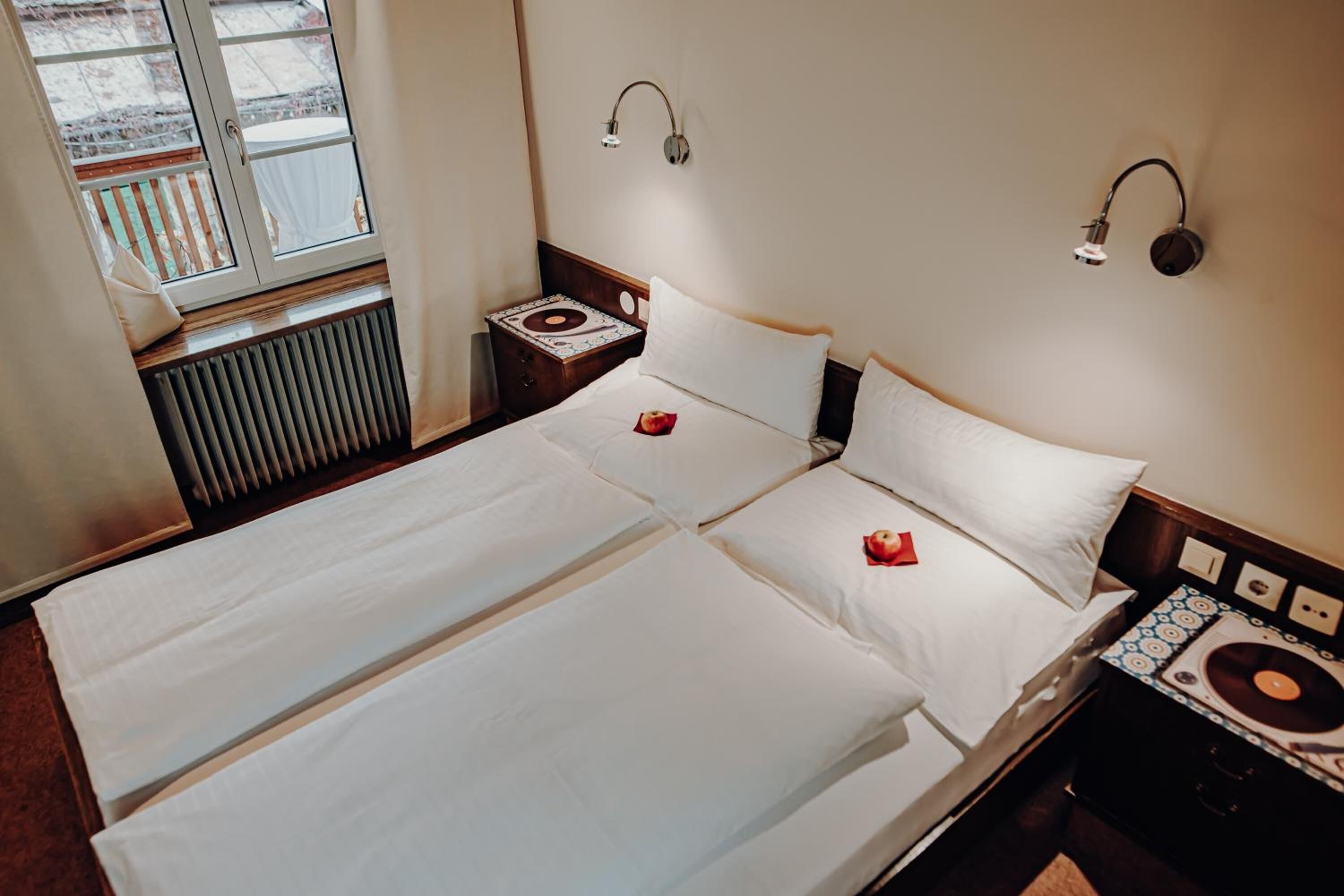 Un lit double avec du linge de lit blanc, deux tables de chevet avec des lampes, un radiateur et un lecteur de disques fixé au mur.