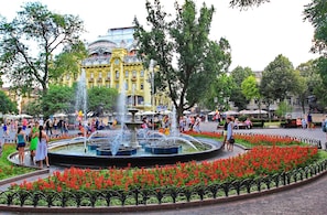 A public square with a central fountain, surrounded by red flowers and trees.