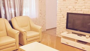 Uma sala de estar com dois poltronas bege, uma mesa de centro branca, uma TV de tela plana em um rack branco, e uma parede com textura de tijolo.