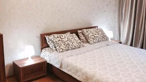 Quarto com cama de madeira, almofadas estampadas, mesa de cabeceira com abajur e papel de parede floral.