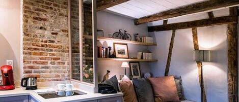 Una habitación acogedora con vigas de madera a la vista, pared de ladrillo, sofá con cojines, cafetera y una lámpara.