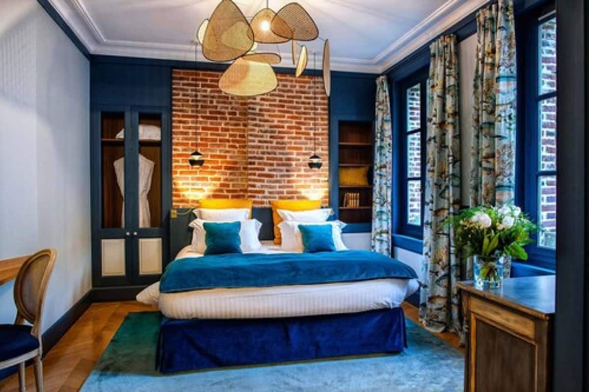 A bedroom with a brick accent wall, a bed with blue bedding, a wooden chair, and a desk with a vase of flowers.