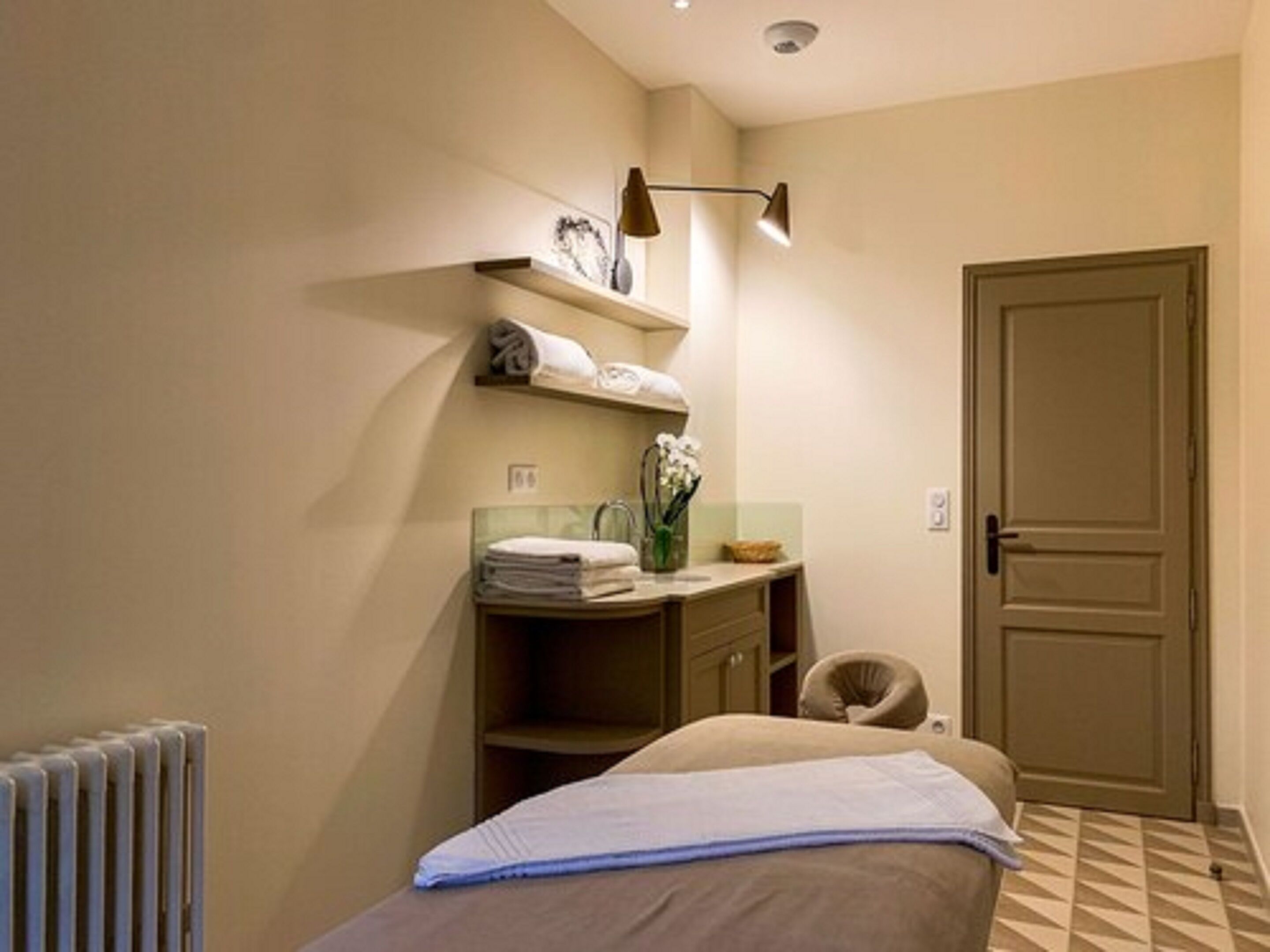Une chambre d’hôtel avec un lit, un radiateur, un bureau avec une chaise, une étagère avec des serviettes et une porte.