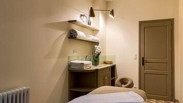 Une chambre d’hôtel avec un lit, un radiateur, un bureau avec une chaise, une étagère avec des serviettes et une porte.