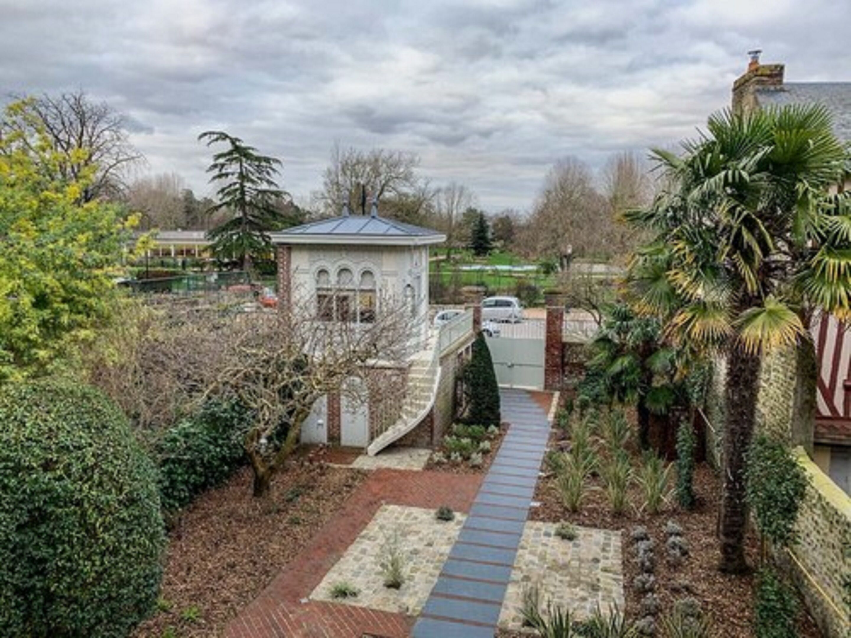 A gated property with a pathway leading to a small white building, surrounded by various plants and trees.