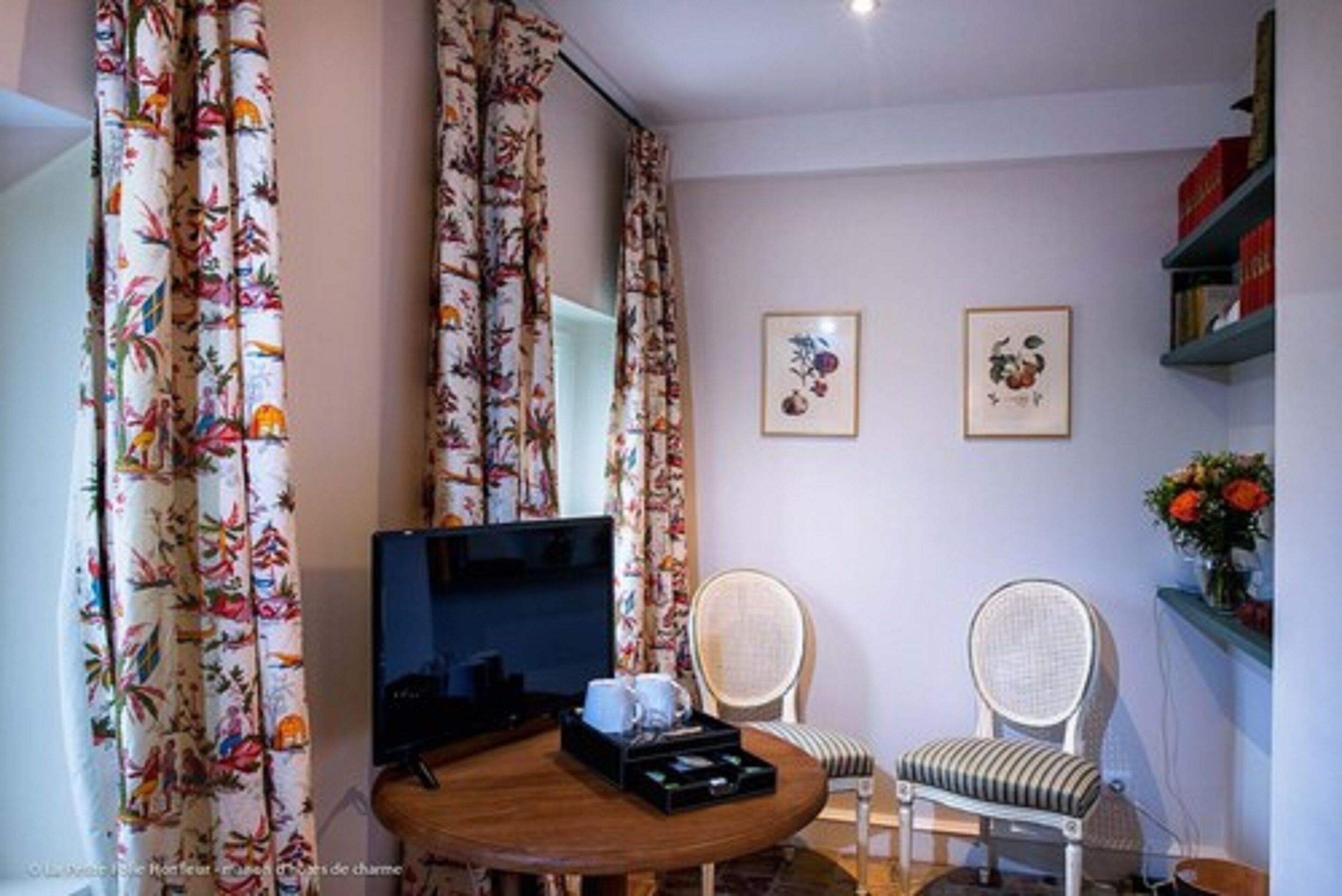 A room with a round wooden table, two wicker chairs, a flat-screen TV, and floral curtains.