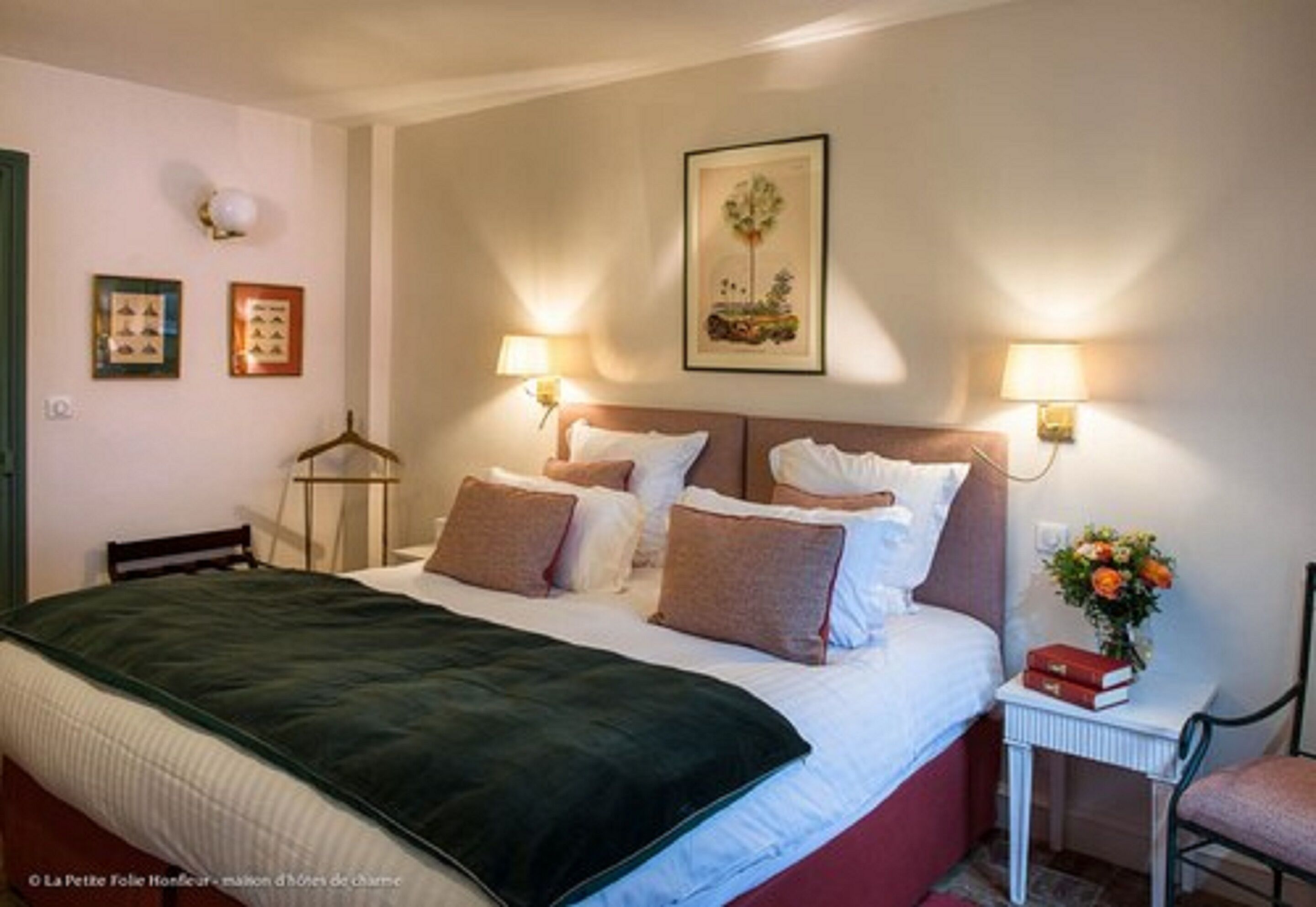 Een net opgemaakt bed met een groene deken, twee nachtlampjes, een ingelijst kunstwerk boven het bed, en een klein tafeltje met een boek en bloemen naast het bed.