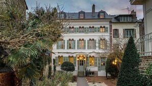 A two-story building with a central entrance, surrounded by a garden with various plants and a pathway leading to the door. - La Petite Folie (Honfleur)