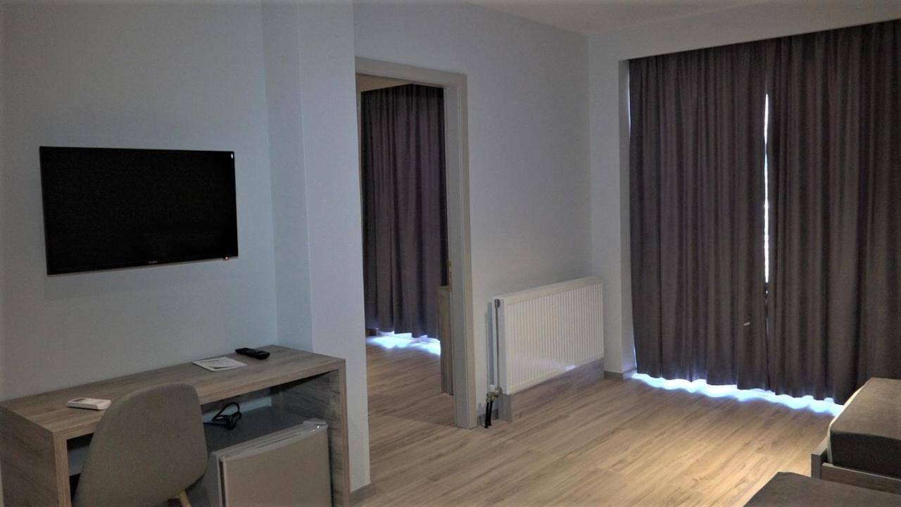 Ein modernes Hotelzimmer mit Schreibtisch aus Holz, Flachbildfernseher, Heizkörper und großen Fenstern mit Vorhängen.