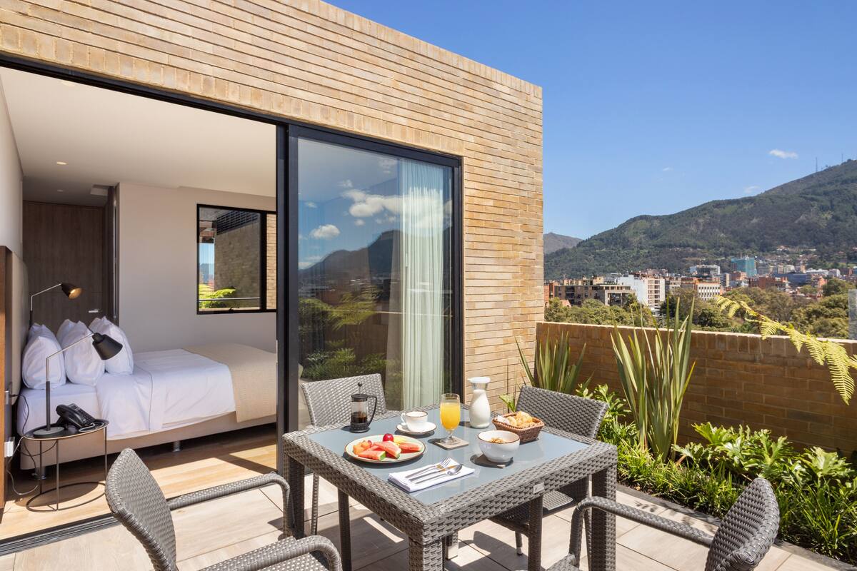 A modern hotel room with a balcony, a bed, a dining table with breakfast, and a city view.