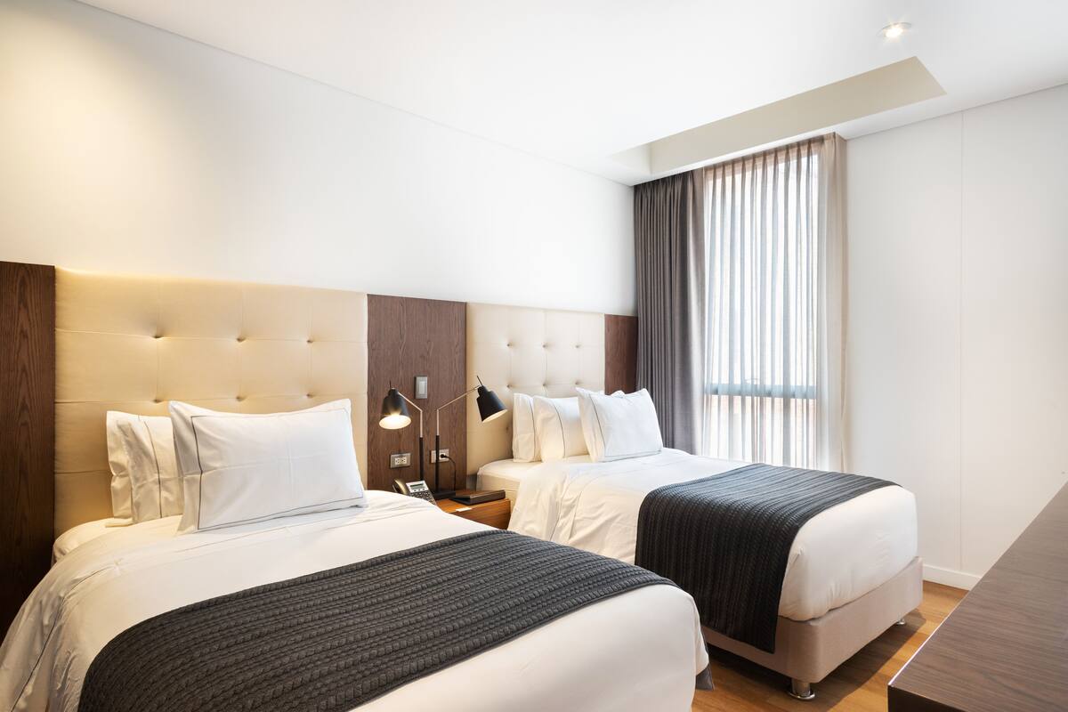 A hotel room with two beds, a wooden headboard, a bedside table with a lamp, and a large window with curtains.