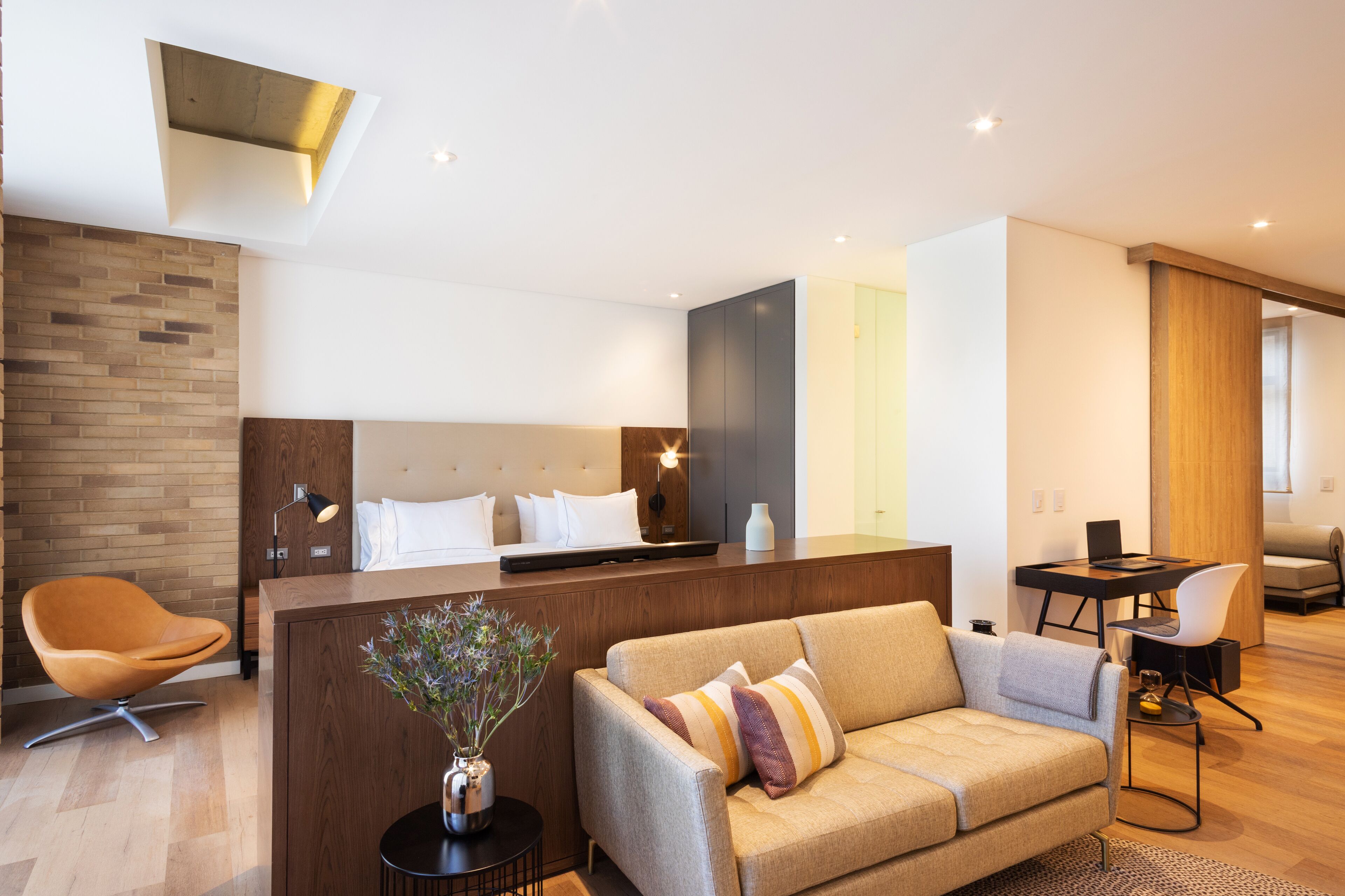 A modern hotel room with a wooden reception desk, a sofa, a dining table, and a large bed with a headboard.