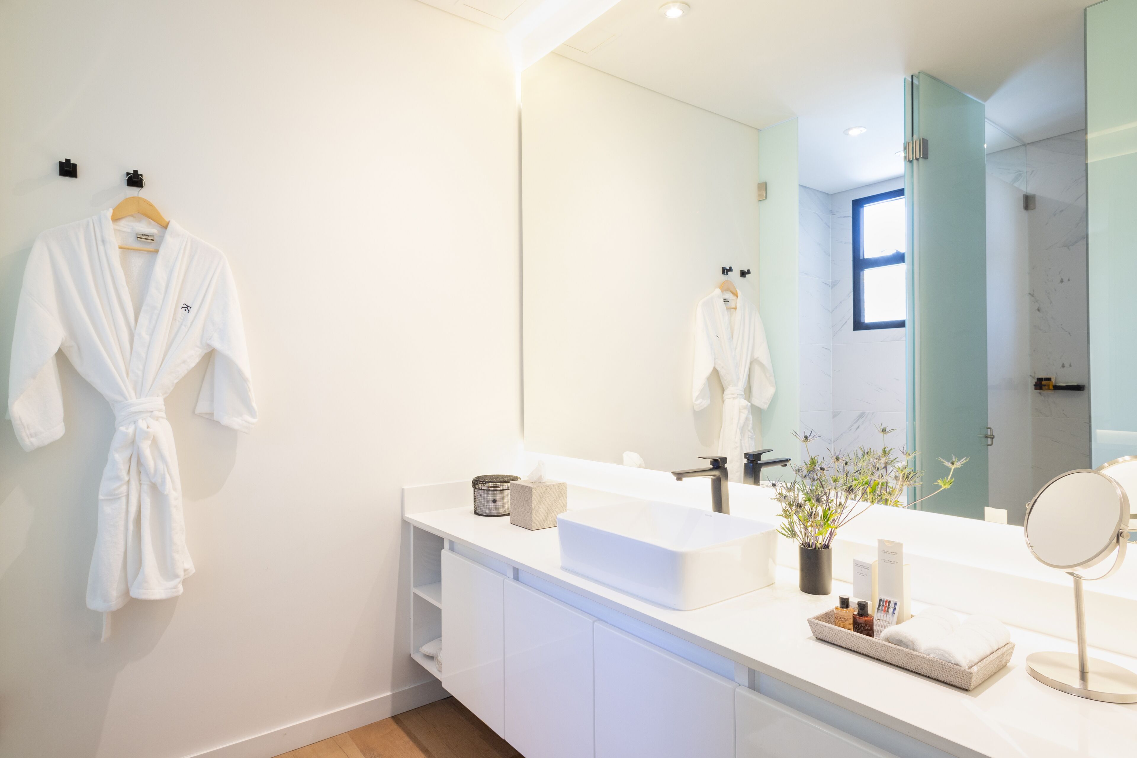 A modern bathroom with a white vanity, a large mirror, and a robe hanging on the wall.