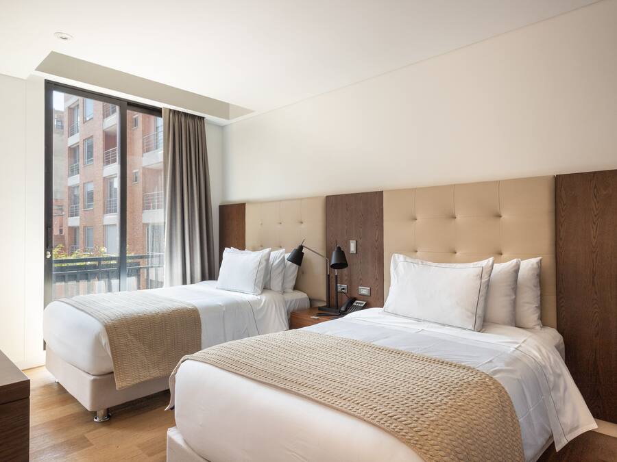 Een hotelkamer met twee bedden, een groot raam met gordijnen, een houten hoofdbord en uitzicht op een gebouw.