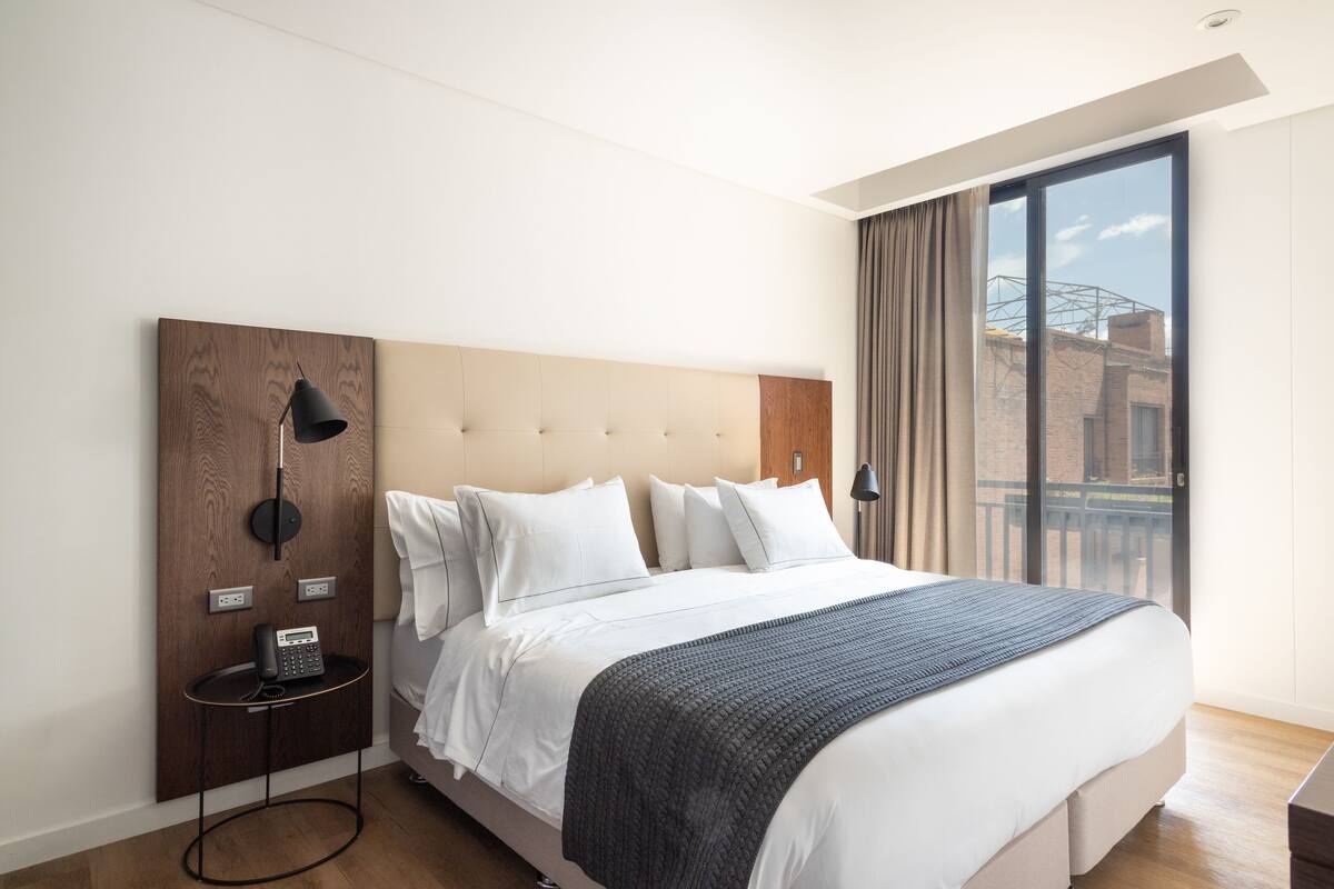 A modern hotel room with a large bed, a wooden headboard, a bedside table with a lamp, and a view of the city through a large window.