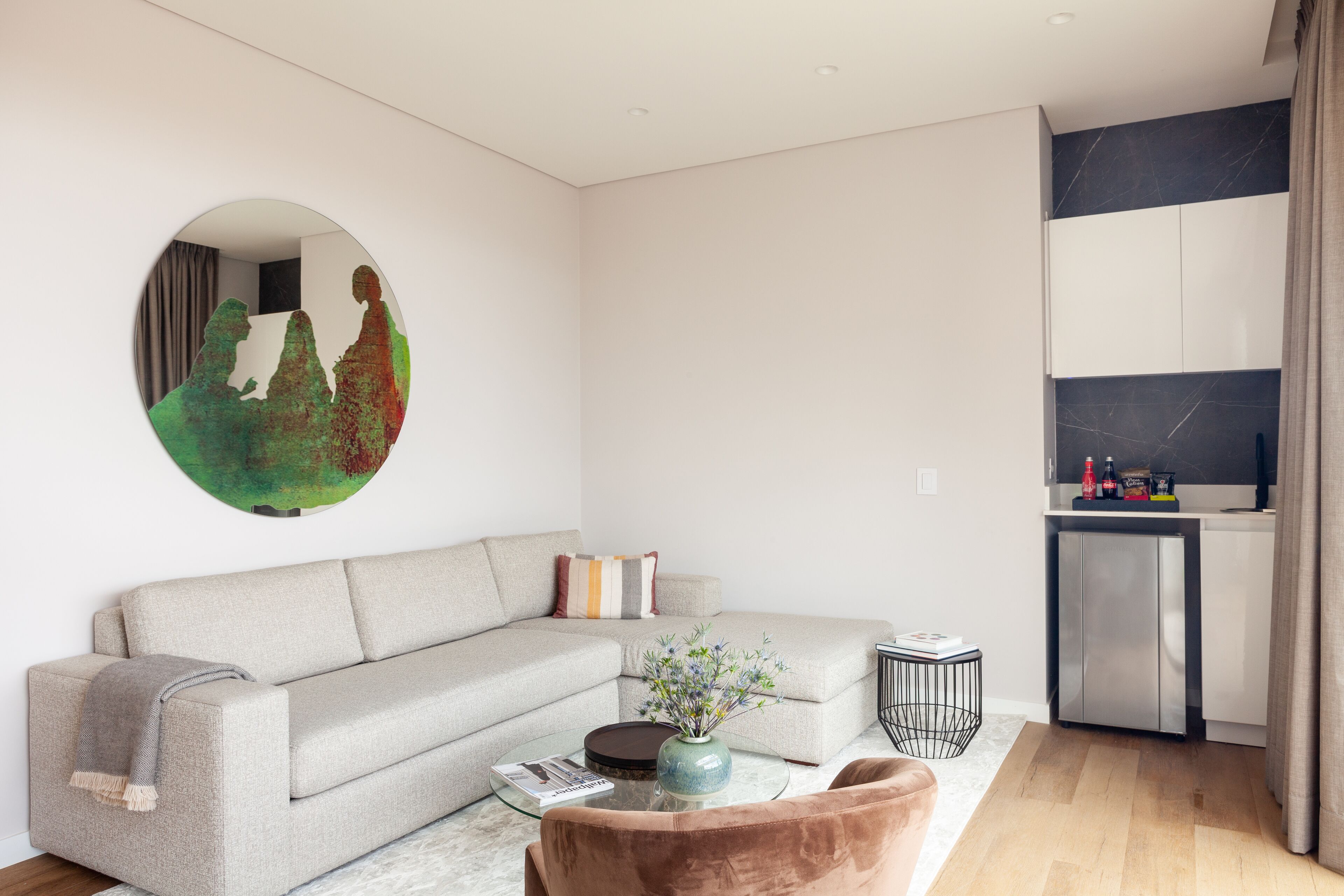 A modern living room with a grey sofa, a round mirror, a coffee table with a vase, and a kitchen area with cabinets and a refrigerator.
