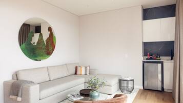 A modern living room with a grey sofa, a round mirror, a coffee table with a vase, and a kitchen area with cabinets and a refrigerator.