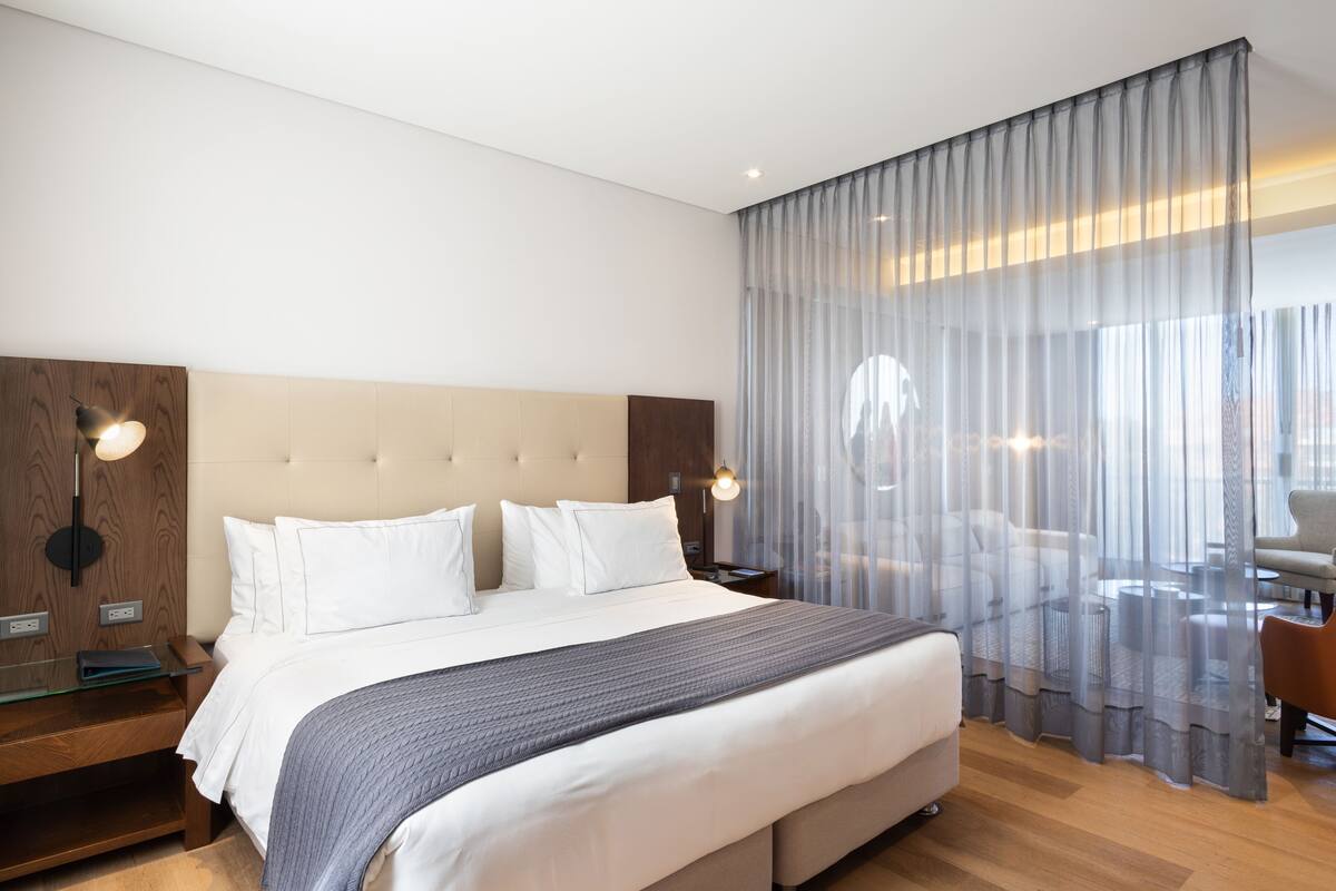 A modern hotel room with a large bed, wooden headboard, bedside tables, and a view through sheer curtains.