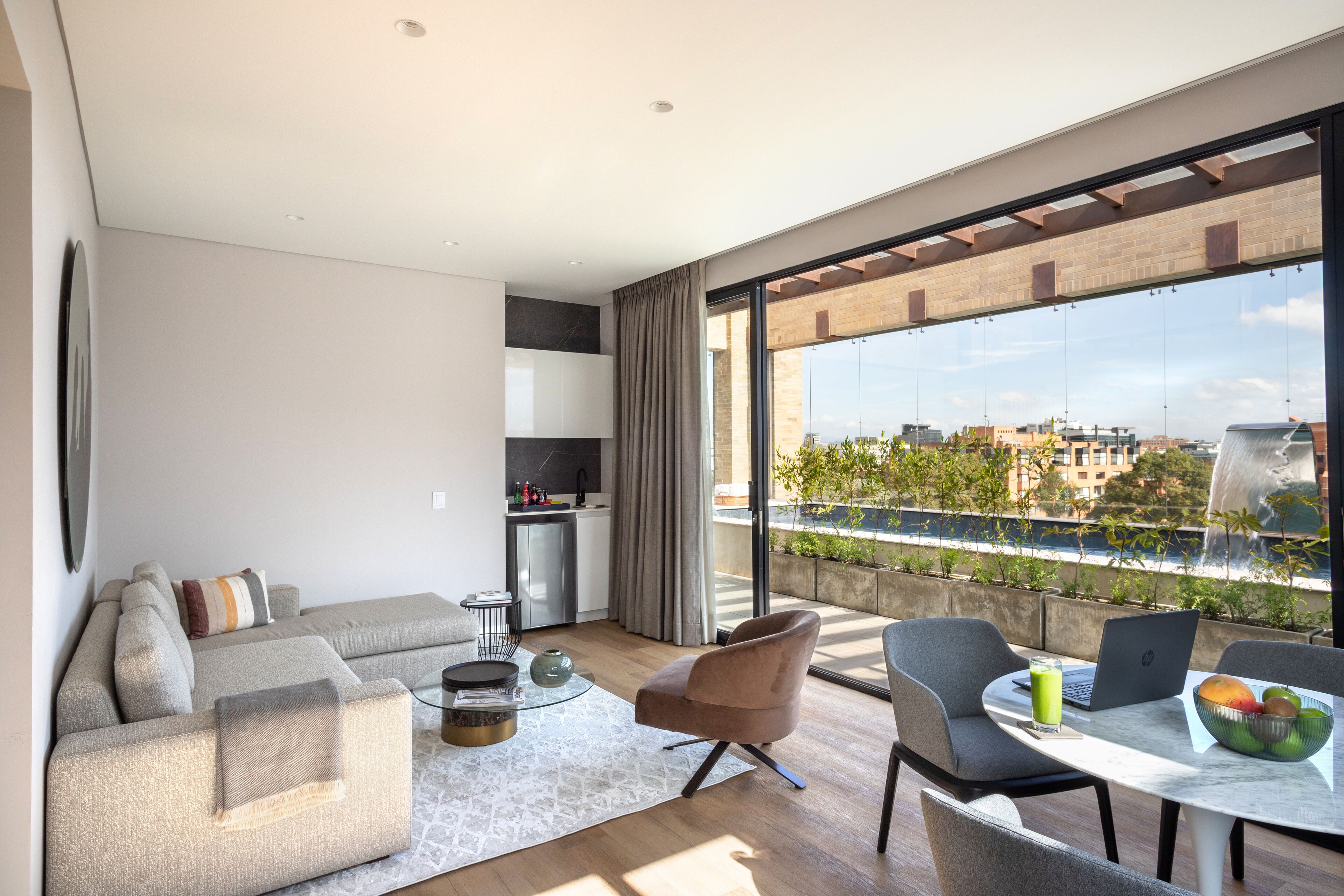 Un salon moderne avec un canapé, des fauteuils, une table à manger sur laquelle se trouvent un ordinateur portable et un panier de fruits, et une vue sur le paysage urbain à travers de grandes fenêtres.