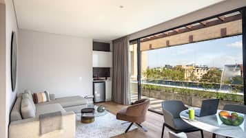 Un salon moderne avec un canapé, des fauteuils, une table à manger sur laquelle se trouvent un ordinateur portable et un panier de fruits, et une vue sur le paysage urbain à travers de grandes fenêtres.