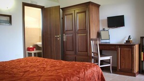A bedroom with a bed, a wooden wardrobe, a desk with a chair, a television, and a small table with a red box.