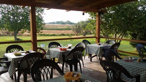 An outdoor patio with tables and chairs, covered by a wooden structure, overlooking a grassy field and trees.