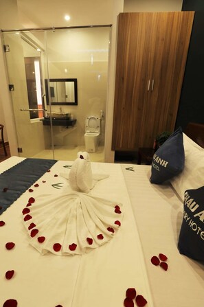 Habitació d'hotel amb un llit decorat amb pètals de rosa i una decoració de cigne blanc, un armari de fusta, un box de dutxa de vidre i un bany visible.