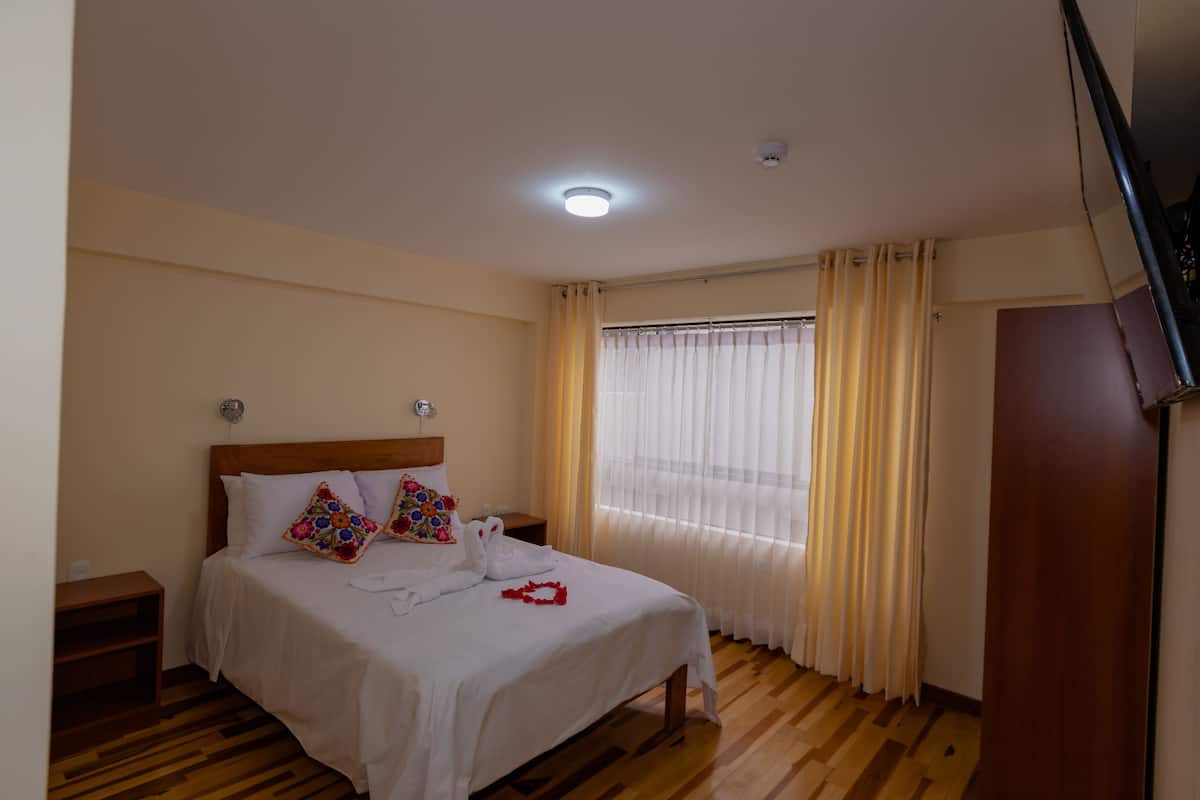 A bedroom with a wooden floor, a bed with white bedding, a nightstand, a window with curtains, and a ceiling light.