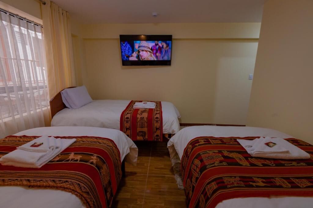 A hotel room with two beds, each with a red and white patterned bedspread and white pillows. A flat-screen TV is mounted on the wall above the beds.