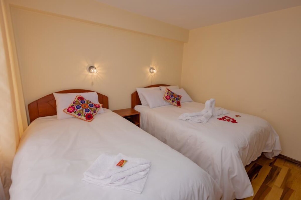 A hotel room with two single beds, each with a white duvet, a floral pillow, and a towel on the bed. There are bedside tables with lamps and a wall-mounted light fixture.