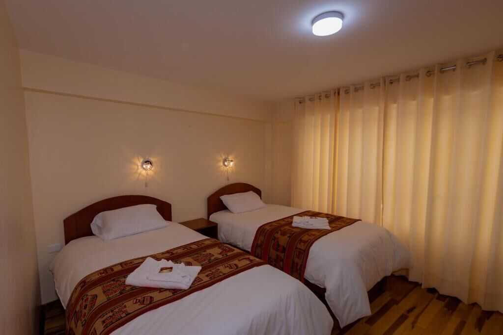Habitación de hotel con dos camas individuales, cada una con una colcha estampada y almohadas blancas. Dos lámparas están fijadas a la pared sobre las camas, y el suelo es de madera.