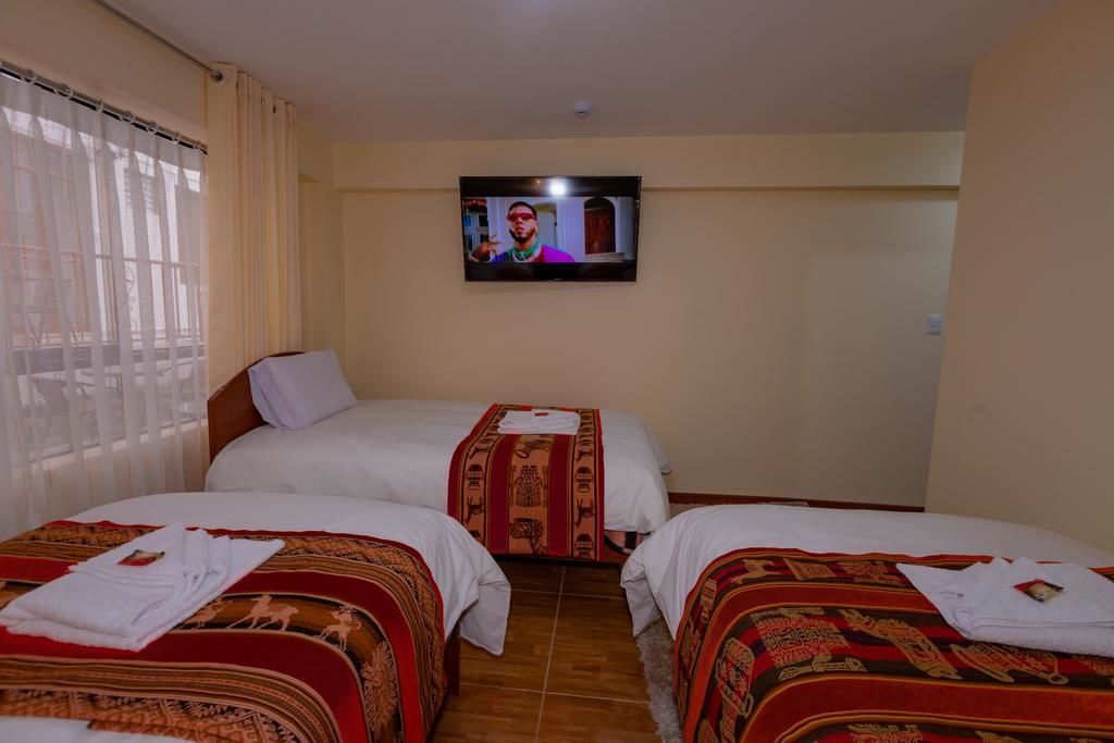 A hotel room with two beds, each with a patterned bedspread and a white pillow. A television is mounted on the wall, and there is a window with curtains.