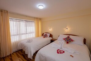 Two single beds with white duvets and decorative pillows, set against a wooden floor and large window with sheer curtains.