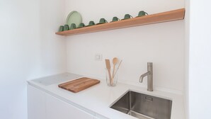 A modern kitchen with a white countertop, a stainless steel sink, a wooden cutting board, and a shelf with green cups. - Masseria Pensato SUITE CARRUBO (Fasano)