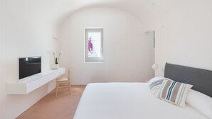 A bedroom with a bed, a TV on a white console, a window with a view of flowers, and a small table with a plant. - Masseria Pensato SUITE CARRUBO (Fasano)
