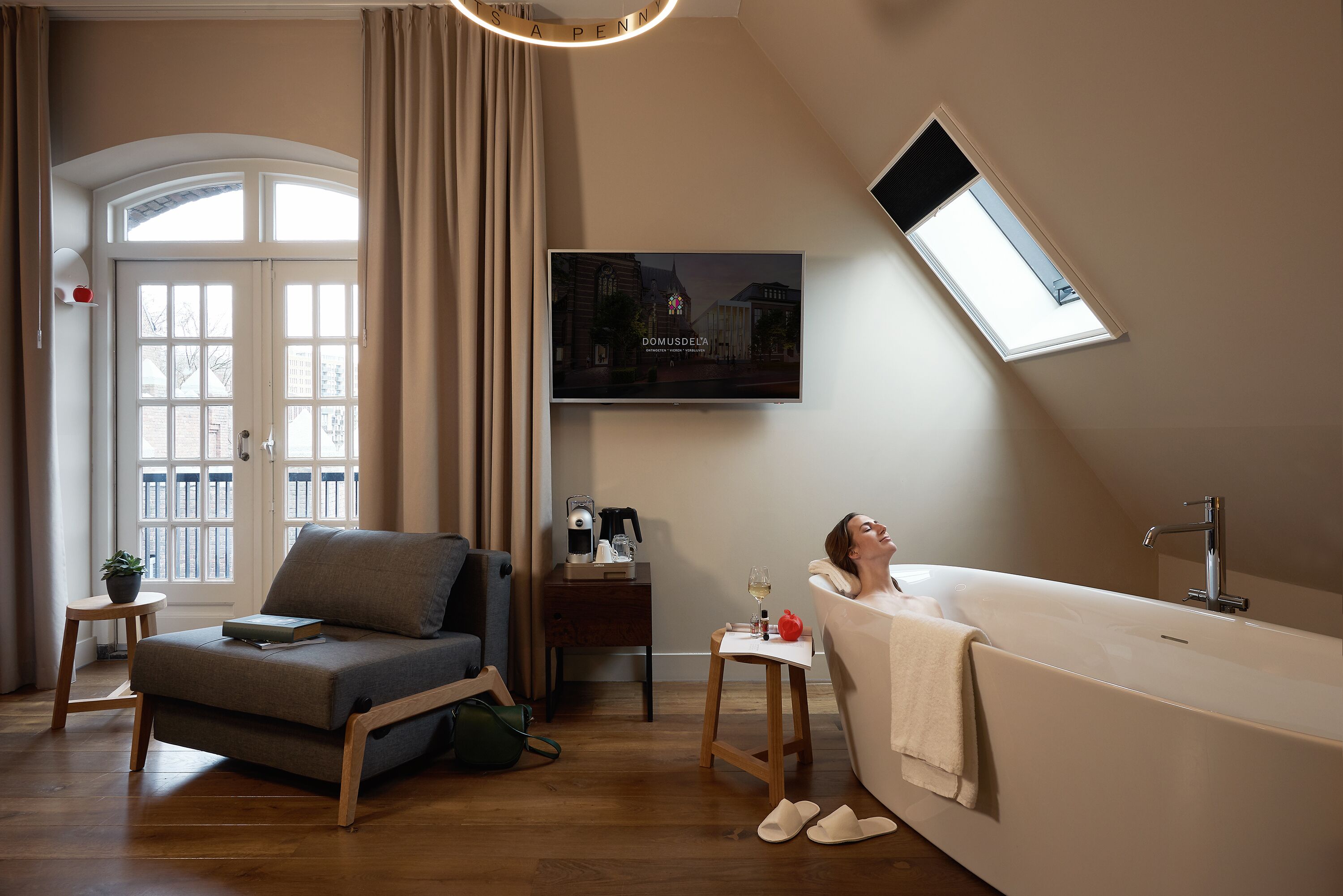 Een moderne badkamer met een badkuip, een aan de muur bevestigde televisie en een raam dat voor daglicht zorgt.