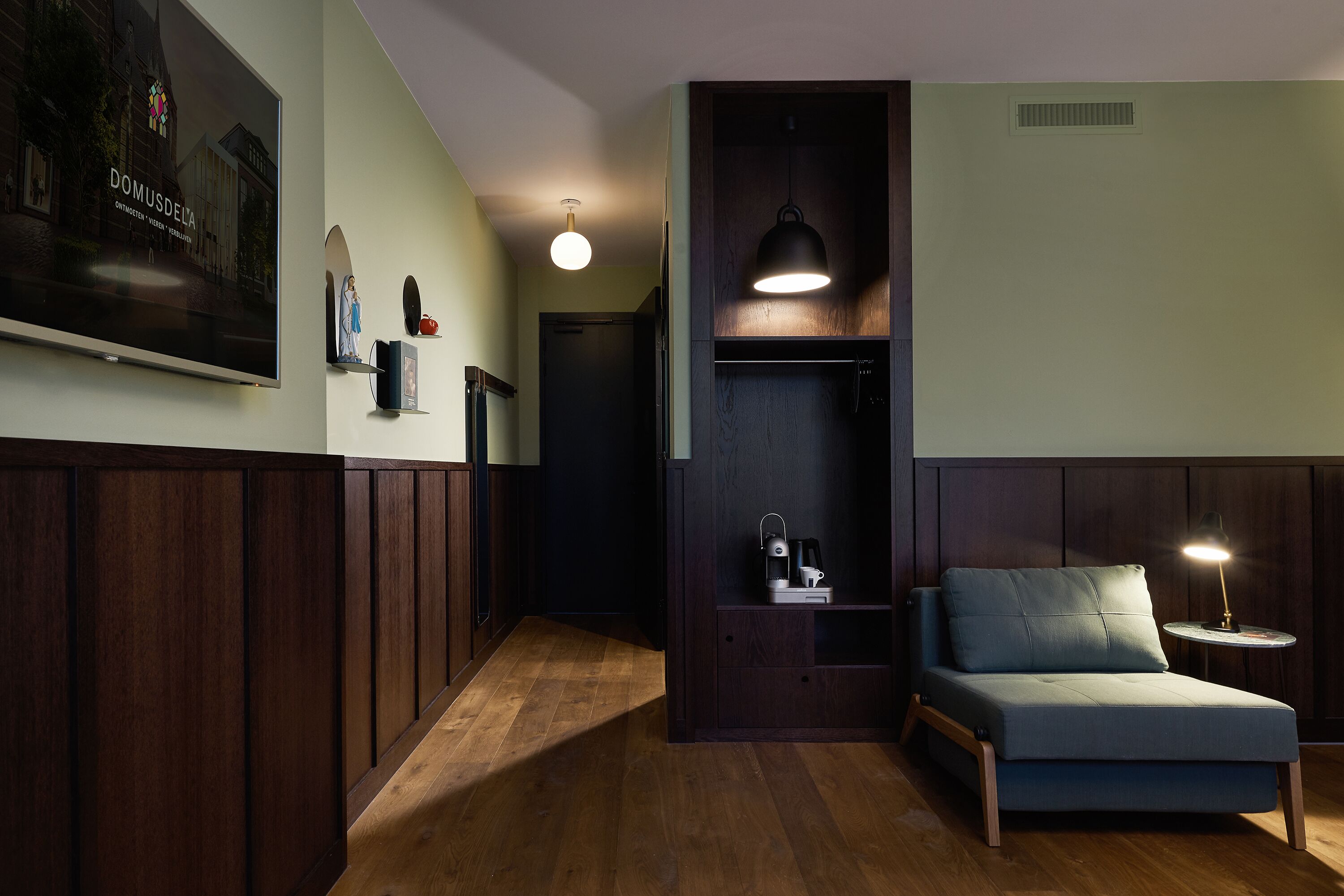 Une pièce moderne avec un plancher en bois, un canapé bleu, une petite table avec une lampe, un téléviseur fixé au mur et une affiche.