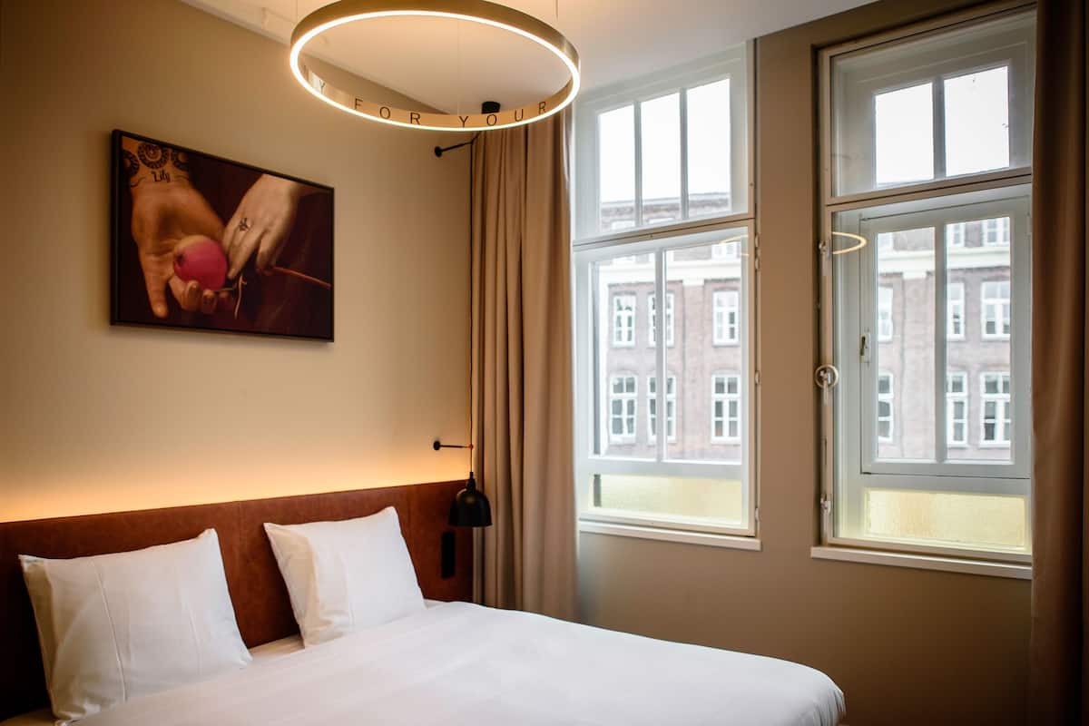 A hotel room with a bed, a painting, a window, and a view of buildings outside.