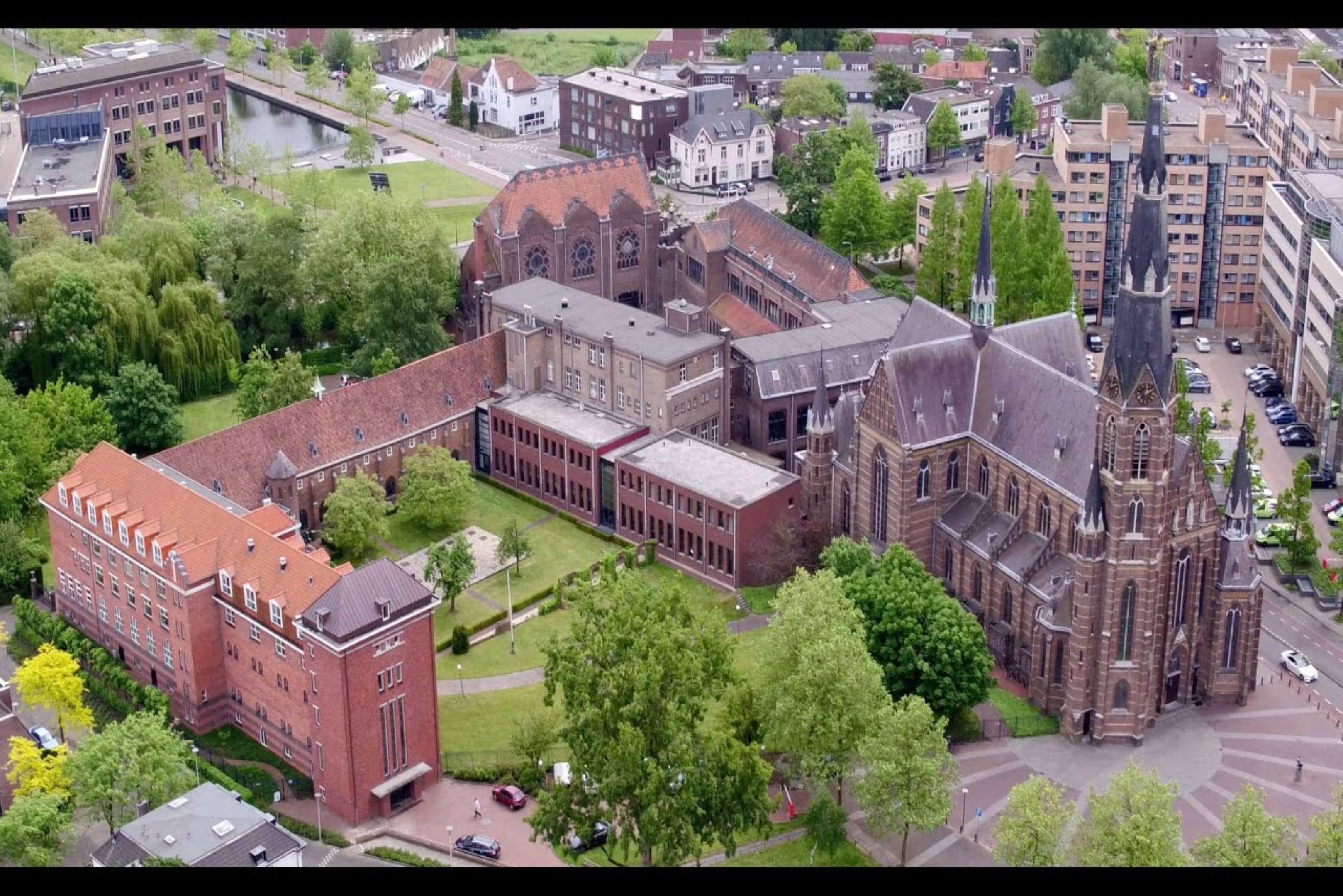 An aerial view of a historic building complex with multiple structures, including a church with a prominent tower, surrounded by green spaces and adjacent buildings.