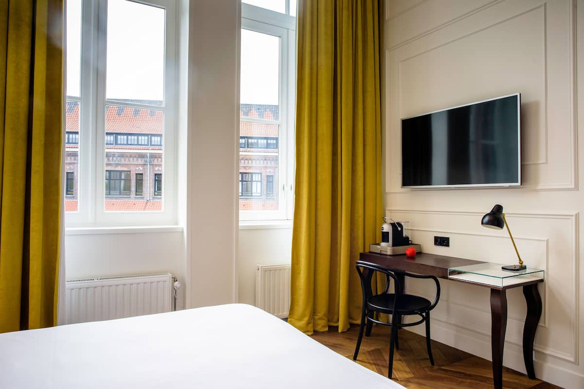 A hotel room with a bed, a desk with a chair, a TV, and a window with curtains.