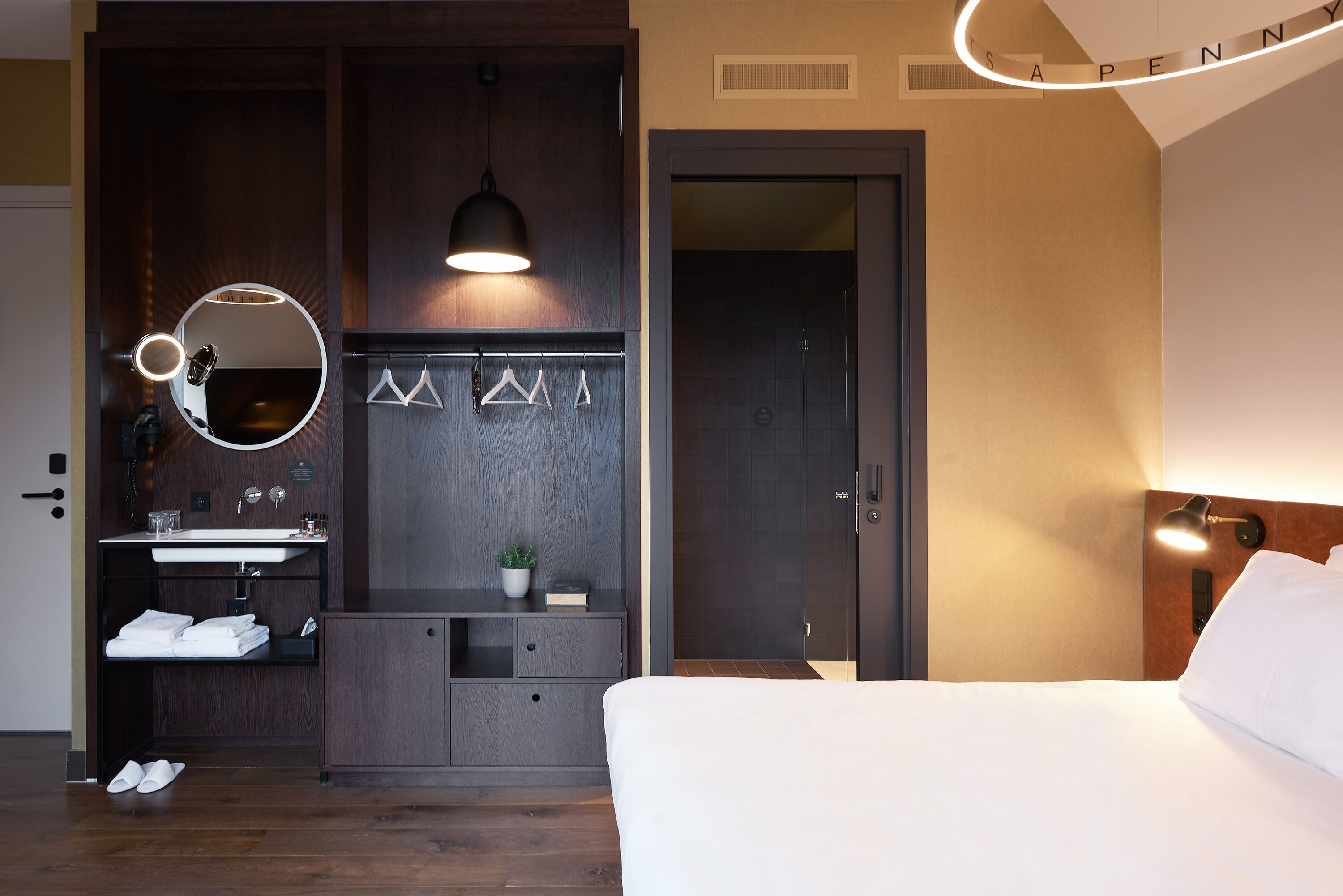 Une chambre d’hôtel comprenant un lit, une table de chevet, un miroir, une lampe suspendue, une armoire et une salle de bain avec un lavabo et un porte-serviettes.