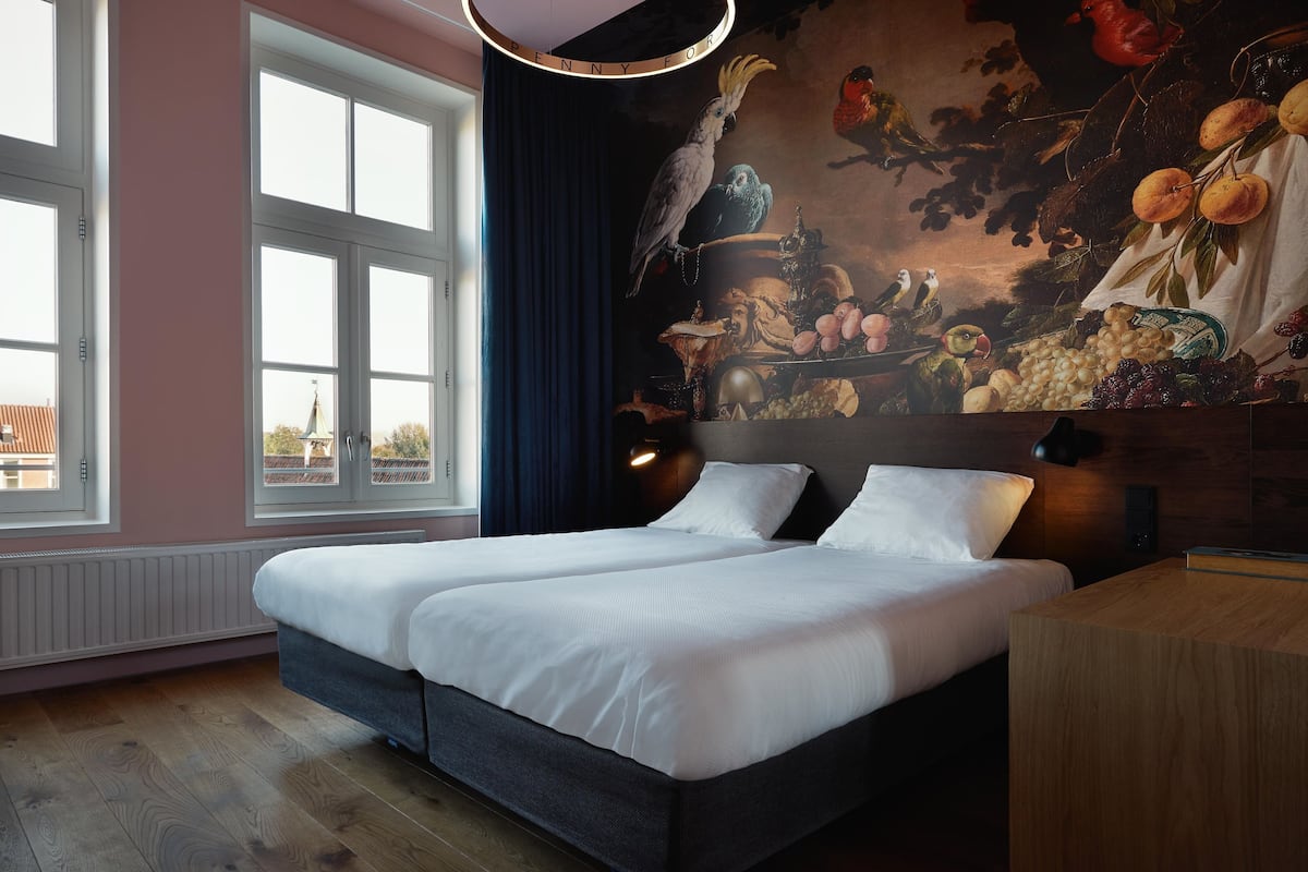 A double bed with white linens, a dark headboard, and a wooden nightstand. A large window with white frames and a view of buildings is visible.