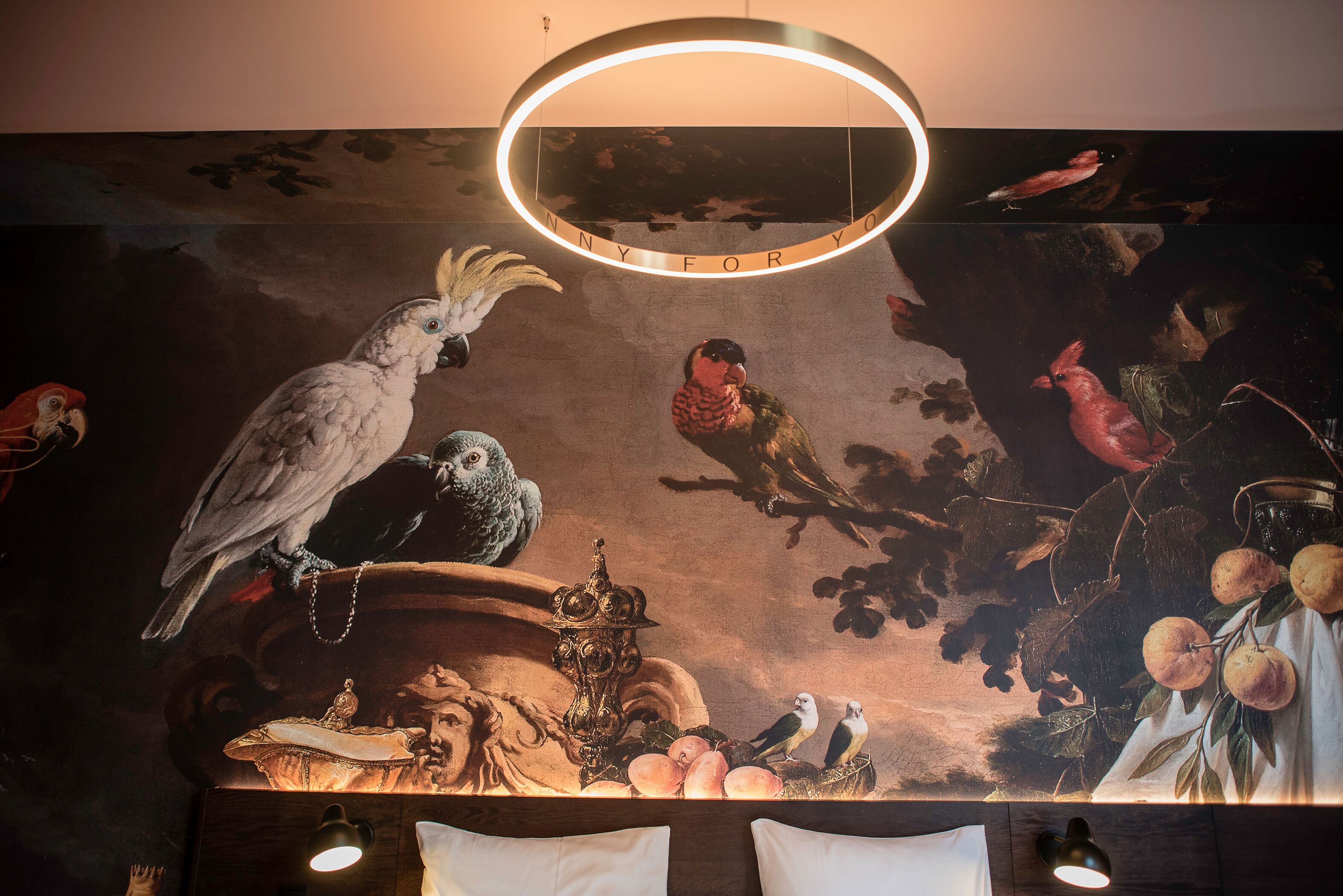 Un mural con aves y fruta, una lámpara circular y una cama con almohadas.