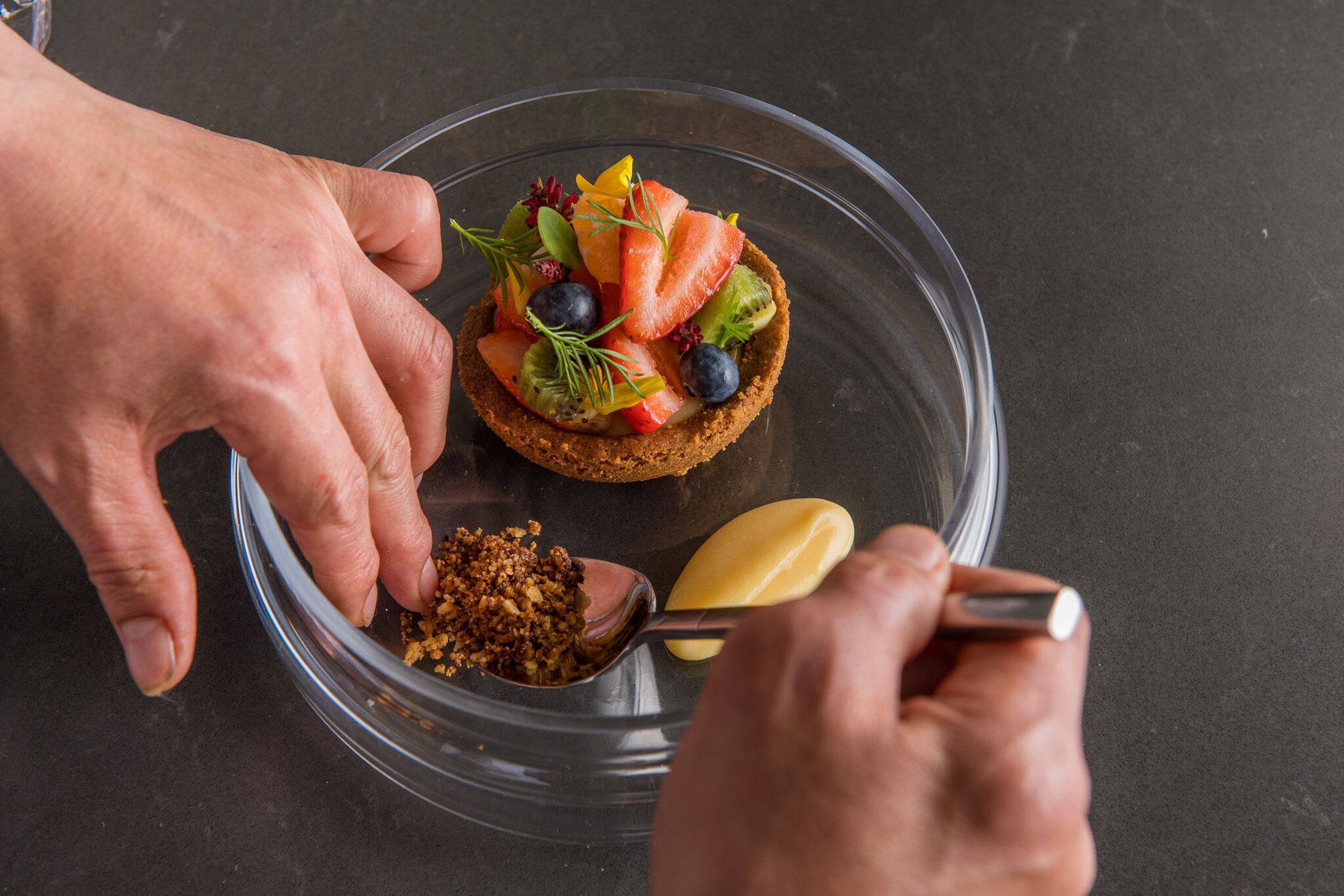 Una persona preparando un postre con un pastel adornado con fruta en un recipiente de vidrio transparente.
