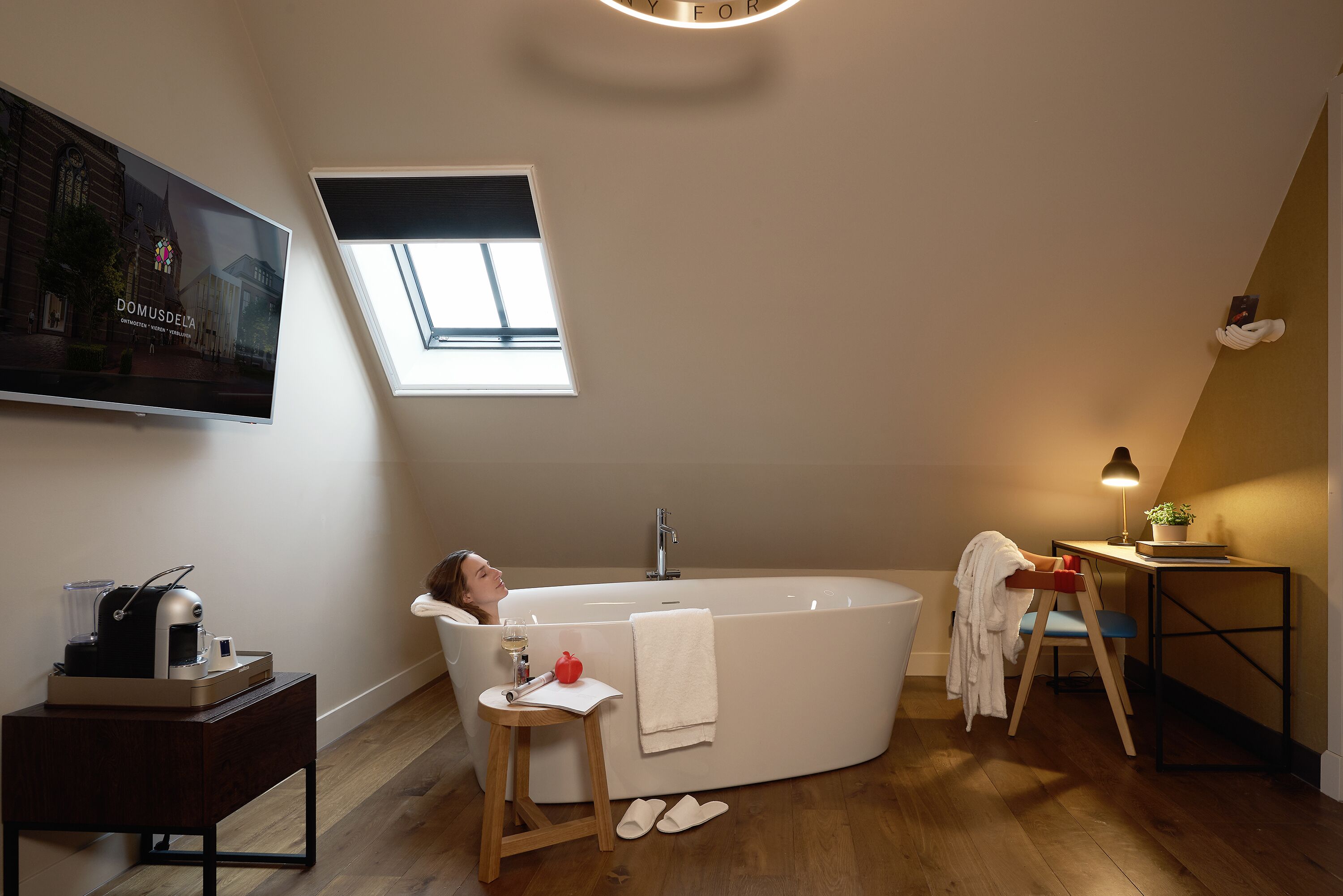 A modern bathroom with a freestanding bathtub, a small table with a lamp, and a window providing natural light.