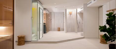 Un couloir d’hôtel moderne avec des portes en verre, un espace douche et une grande plante en pot.